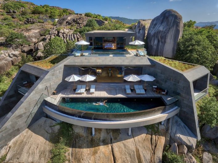 Luxuriöse Villa auf felsigem Gelände mit drei Ebenen: Terrasse mit Infinity-Pool und schwimmender Person unten, mittlere Ebene mit Liegestühlen und Sonnenschirmen, oben ein weiterer Pool mit Liegeflächen.