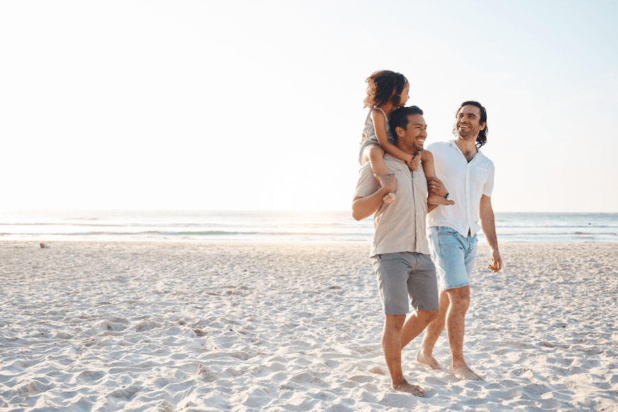Zwei Männer spazieren am Strand, einer trägt ein Mädchen auf den Schultern. Der Himmel ist klar, das Meer im Hintergrund. Sie lachen und genießen den sonnigen Tag.