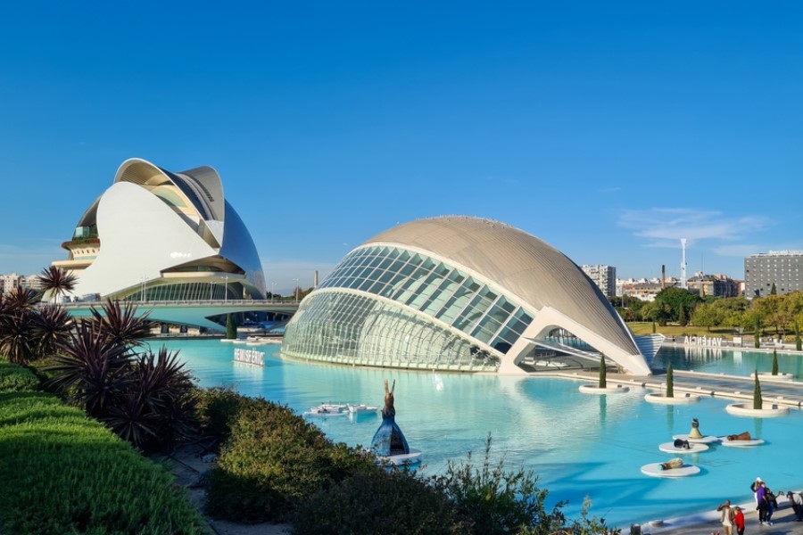 Moderne weiße Gebäude mit geschwungenen Formen im Wasser, umgeben von blauer Lagune und Pflanzen im Vordergrund, strahlend unter blauem Himmel, im Hintergrund eine Stadtansicht.