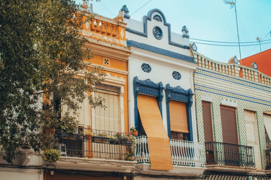 Bunte, reich verzierte Fassaden von drei Reihenhäusern in El Cabanyal, Valencia. Links orangefarben, in der Mitte blau und weiß, rechts grün-weiß kariert mit Fliesen. Bäume am linken Bildrand.