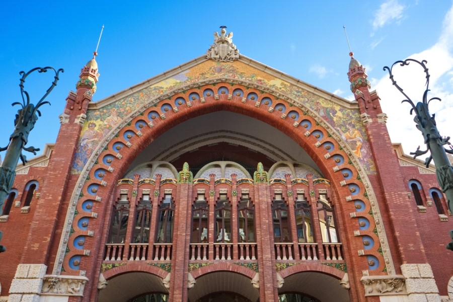 Bogenförmiger Eingang des Mercado de Colón in Valencia aus rotem Backstein mit kunstvollen Mosaiken verziert. Zwei Türme flankieren den Eingang, bei blauem Himmel am Tag.