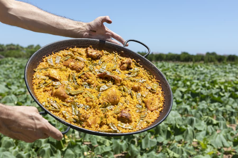 Große Paella-Pfanne mit Safranreis, Hühnchen und grünen Bohnen wird im Vordergrund von Händen gehalten. Im Hintergrund breitet sich ein grünes Feld unter blauem Himmel aus.