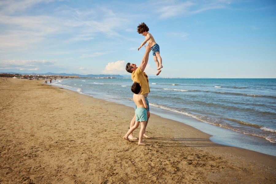 Ein Mann hebt ein Kind freudig am Strand hoch, während ein anderes Kind neben ihm steht. Der Sandstrand erstreckt sich nach links, das blaue Meer mit kleinen Wellen rechts. Klarer Himmel.