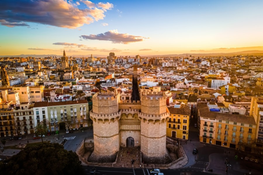 Im Vordergrund stehen die Torres de Serranos, zwei große, alte Türme aus Stein. Dahinter erstreckt sich eine Stadtlandschaft unter einem goldenen Sonnenuntergang mit vereinzelten Wolken.